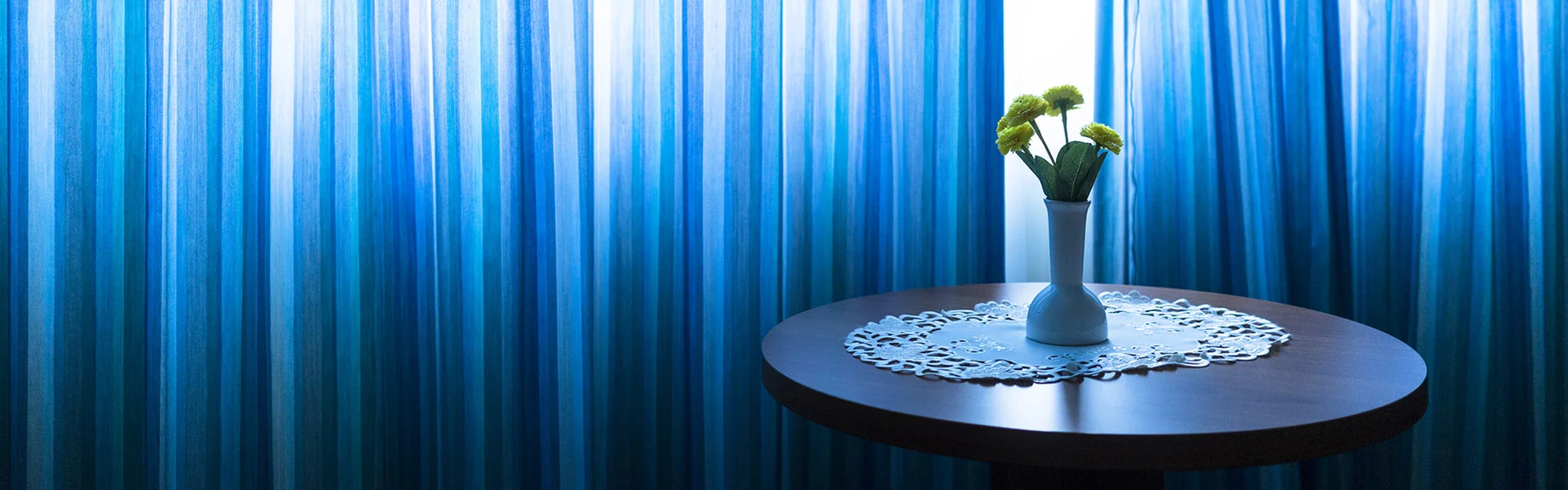 Reportagefotografie: Runder Tisch mit Blumenvase vor einem blauen Vorhang in einem Hotelzimmer.