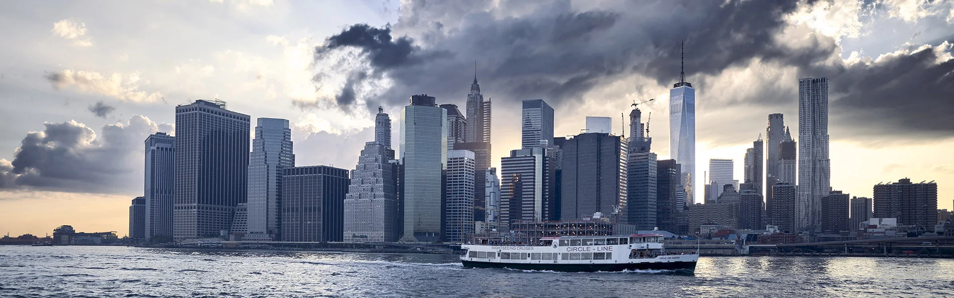 Reportagefotografie: Skyline von Manhattan mit dem One World Trade Center am Wasser unter dunklen Wolken.